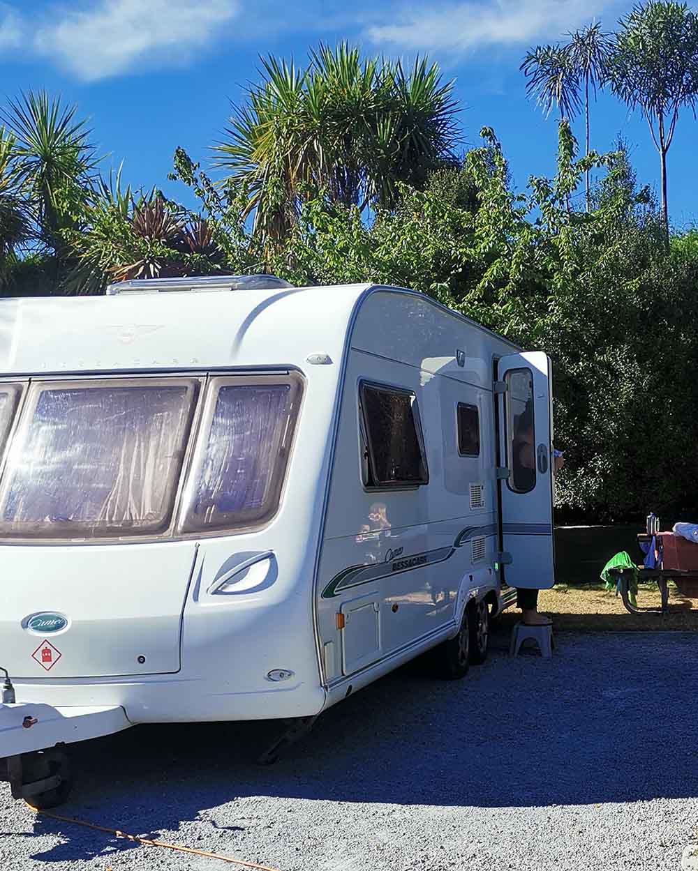White caravan parked on gravel at NZ campground with doors open and camping gear