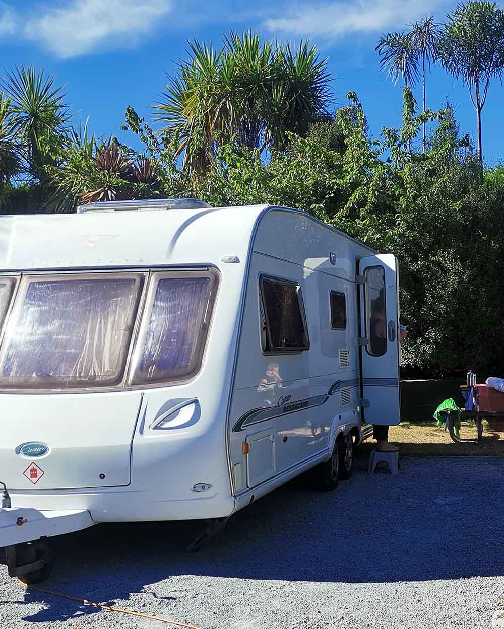 White caravan parked on gravel at NZ campground with doors open and camping gear 