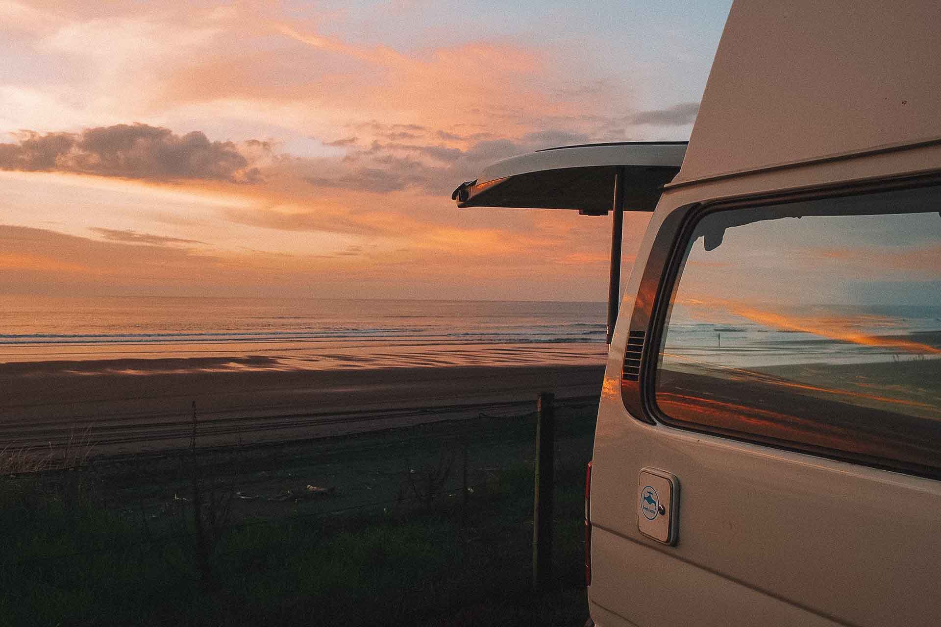 Beach in NZ at sunset with caravan parked and boot open facing pink clouds